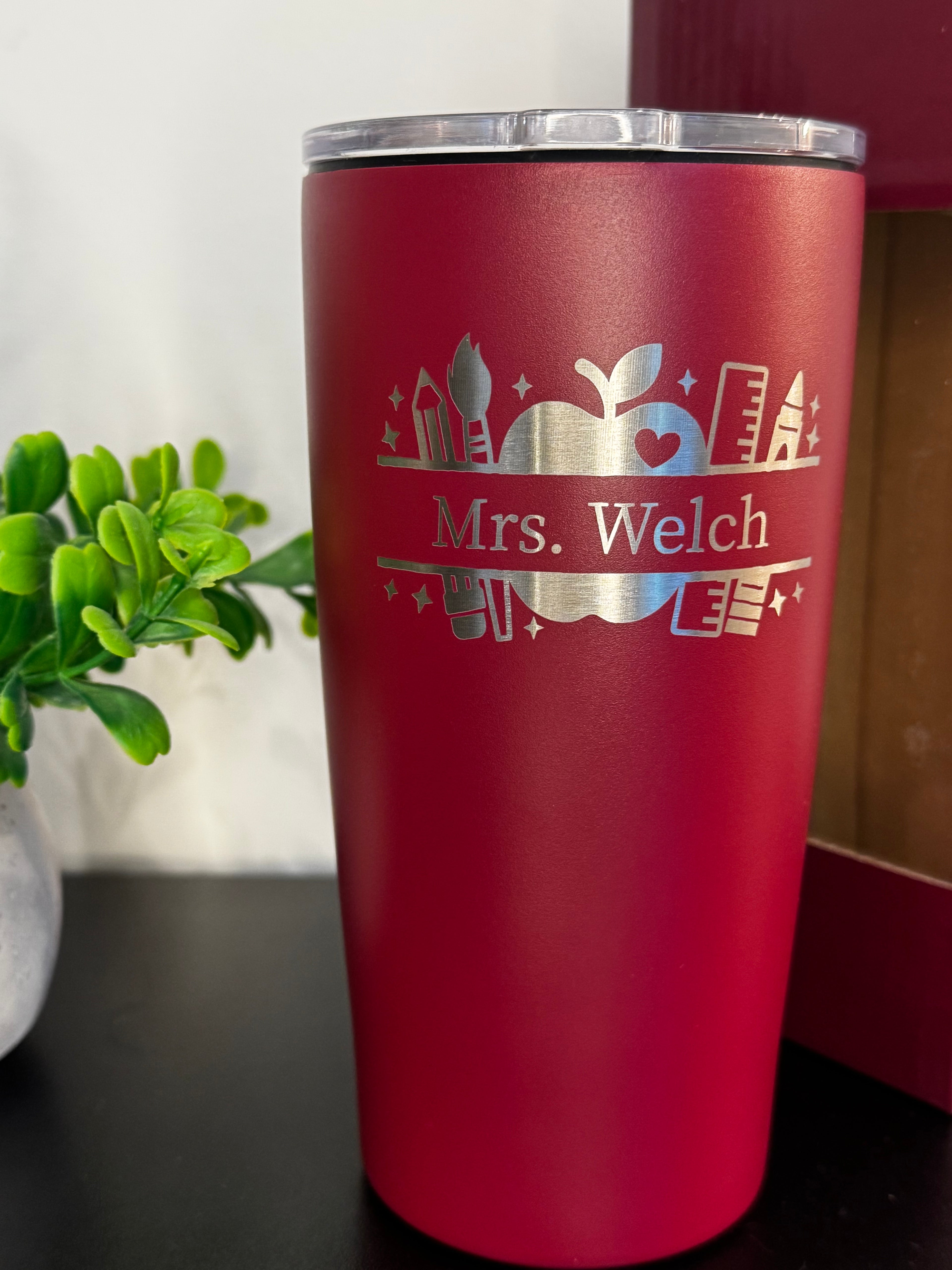 A maroon insulated tumbler with a silver lid, featuring a silver laser-engraved design of an apple, books, and the text 'Mrs. Welch', shown on a dark tabletop with a blurred green plant in the background.