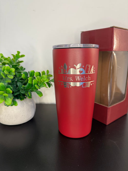 A maroon insulated travel tumbler with a silver lid stands on a dark tabletop, next to a small potted green plant and a red gift box with a clear window. The tumbler is printed with a silver holiday design and the name 'Mrs.