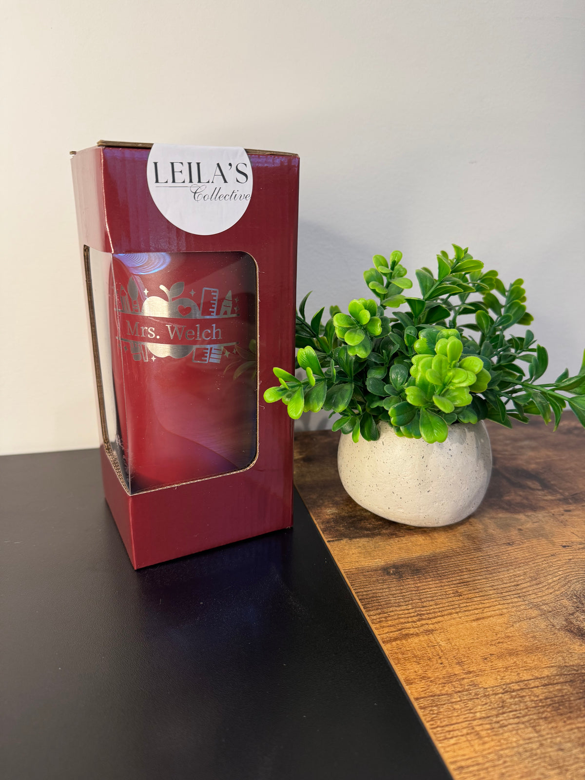 A red glass candle in a clear window box package from Leila's Collection, placed on a dark tabletop next to a small green potted plant against a light wall.