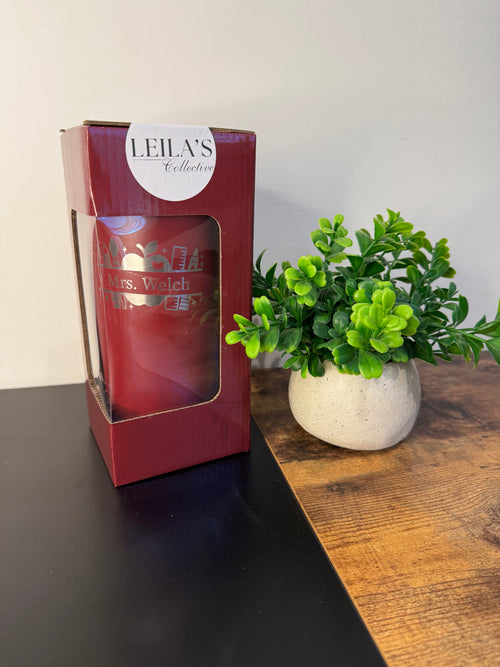 A red glass candle in a clear window box package from Leila's Collection, placed on a dark tabletop next to a small green potted plant against a light wall.