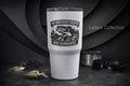 A tall white insulated tumbler with a black fish graphic and the text 'MY BROTHER'S KEEPER / BASS TOURNAMENT' stands on a dark tabletop. Fishing lures, a spool of line, and sunglasses are arranged around it, with a dark textured backdrop.