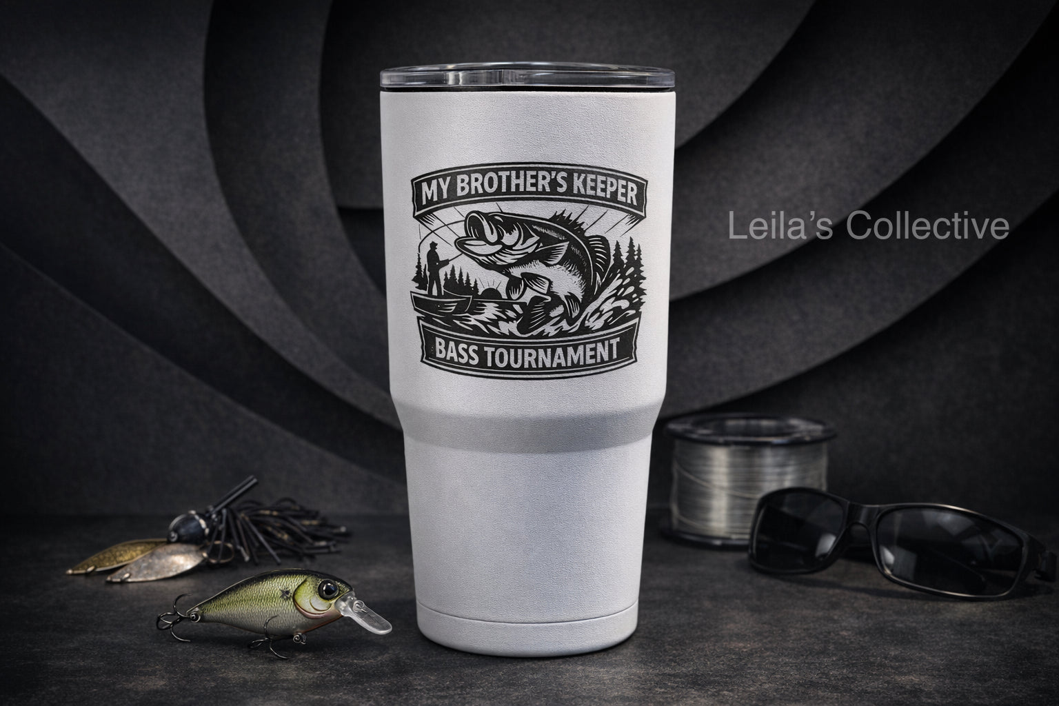A tall white insulated tumbler with a black fish graphic and the text 'MY BROTHER'S KEEPER / BASS TOURNAMENT' stands on a dark tabletop. Fishing lures, a spool of line, and sunglasses are arranged around it, with a dark textured backdrop.