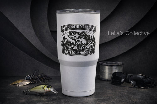 A tall white insulated tumbler with a black fish graphic and the text 'MY BROTHER'S KEEPER / BASS TOURNAMENT' stands on a dark tabletop. Fishing lures, a spool of line, and sunglasses are arranged around it, with a dark textured backdrop.