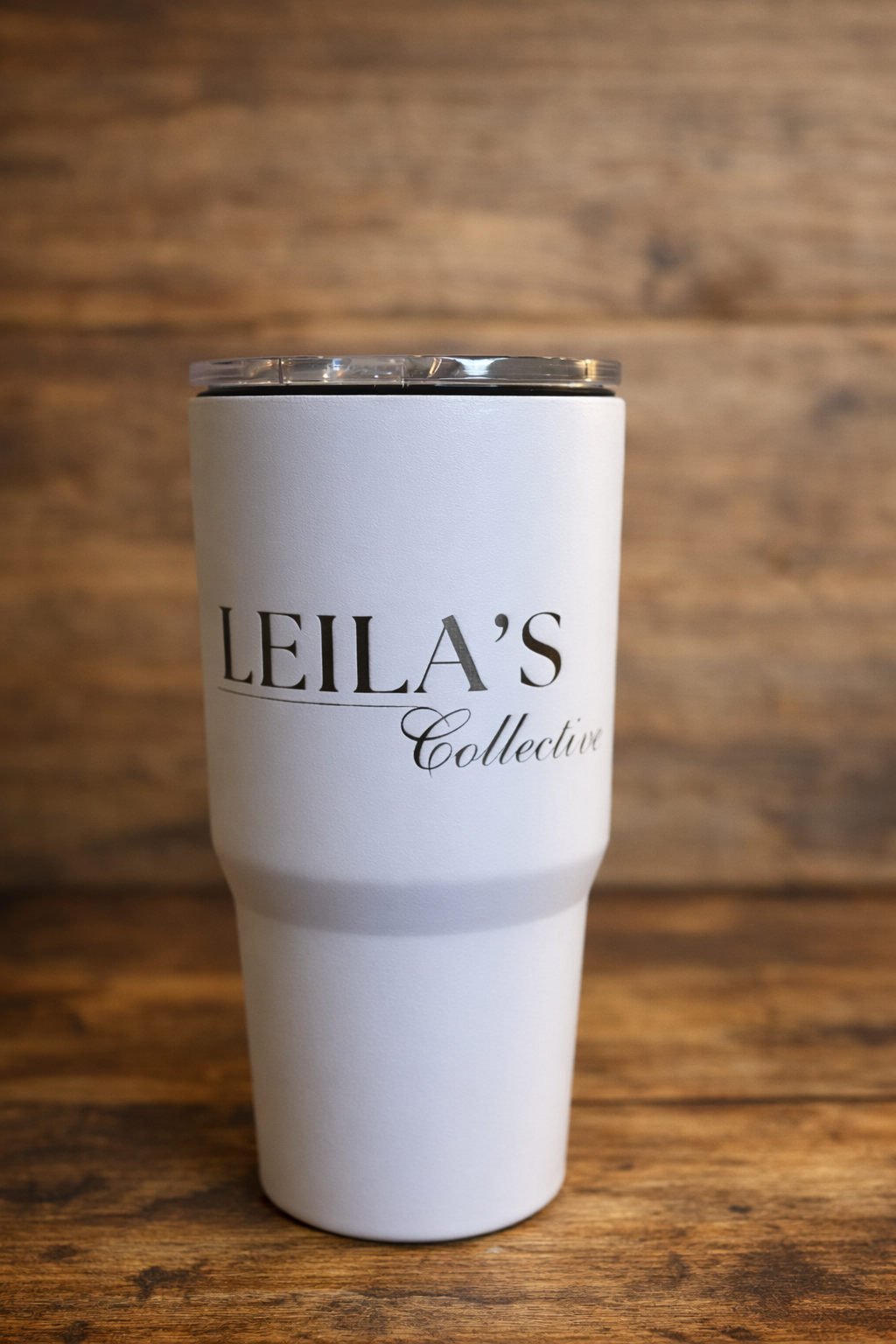 A white insulated tumbler with a clear lid, printed with the text 'LEILA'S Collective' in black, set against a rustic wooden background.