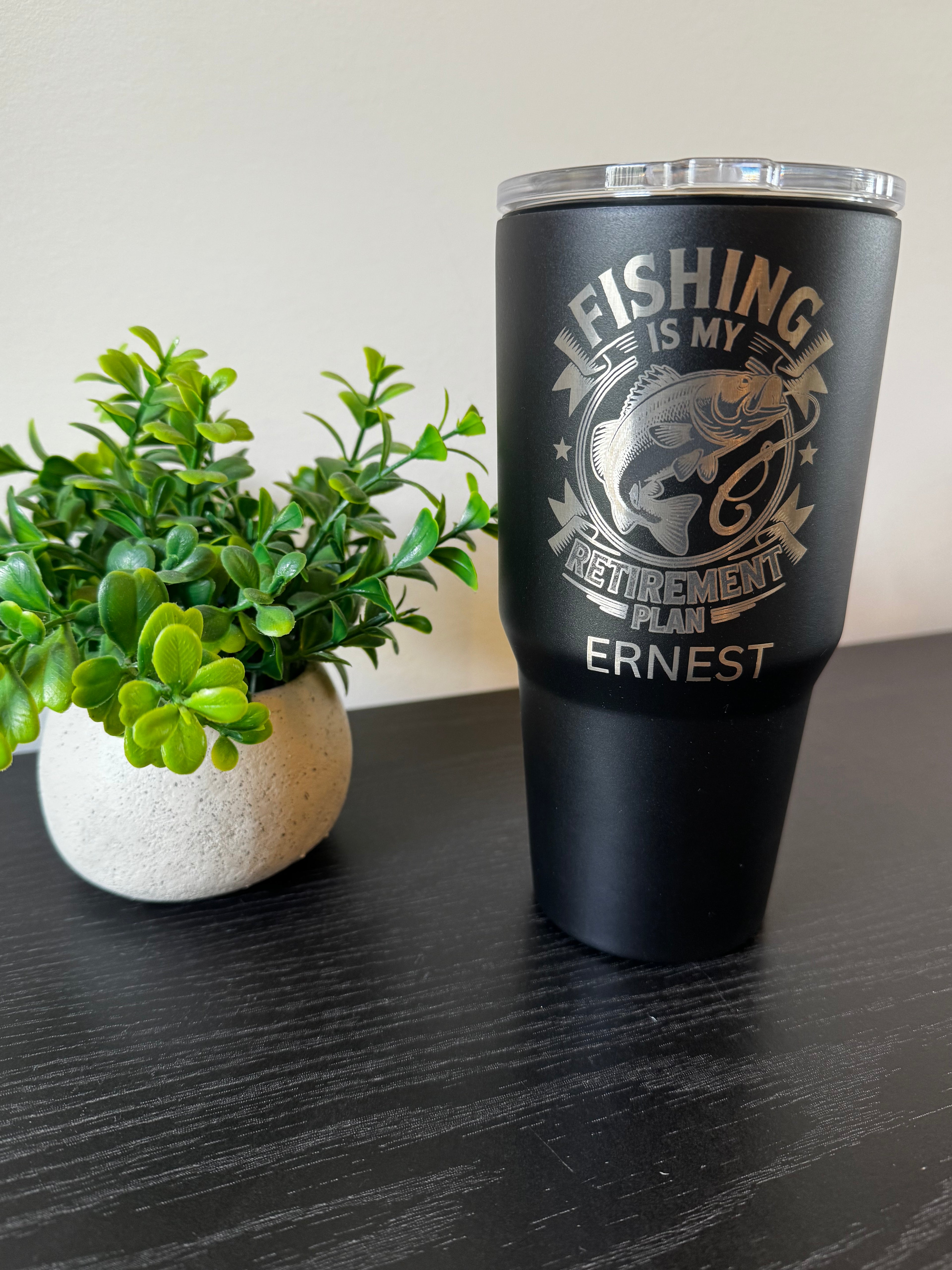 A matte black insulated tumbler with a clear lid stands on a dark tabletop next to a small green potted plant. The tumbler is printed with a silver fish graphic and the text “FISHING IS MY RETIREMENT PLAN ERNEST.