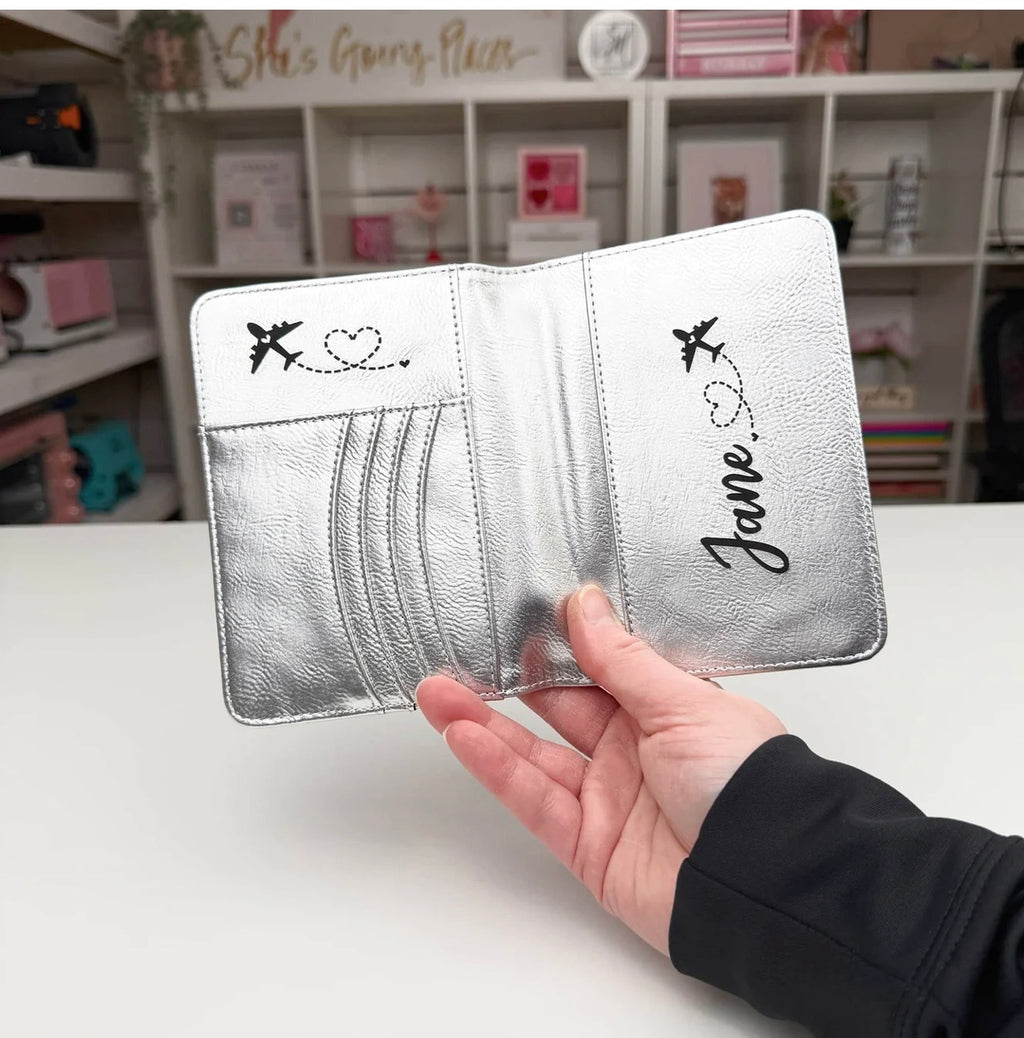 Silver passport holder with 'Jane' engraved, held by a hand on a white surface.
