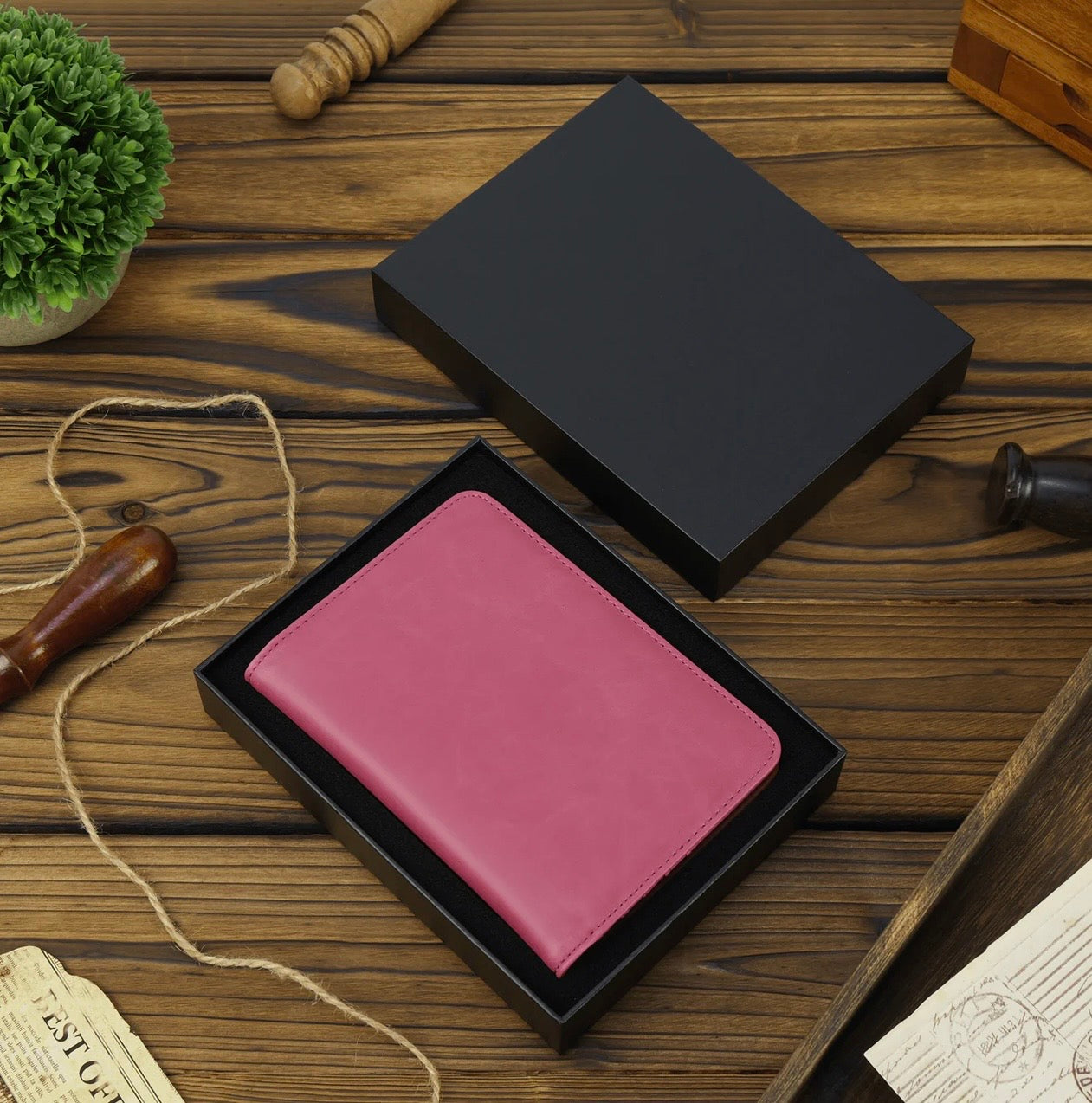Pink leather passport wallet in a black box on a wooden surface with decorative items.