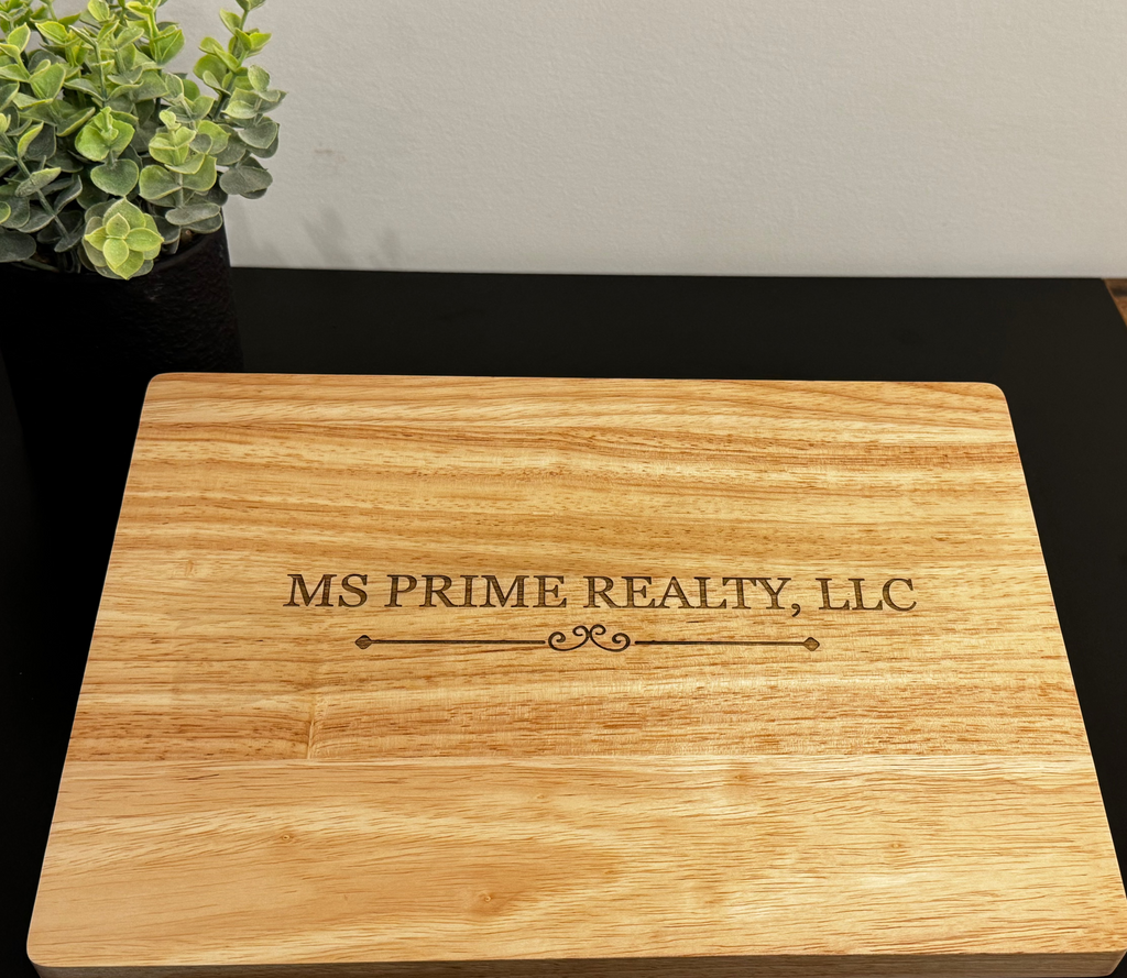 A rectangular wooden cutting board with natural grain, featuring the engraved text 'MS PRIME REALTY, LLC' in the center, resting on a black tabletop with a small potted plant in the background.