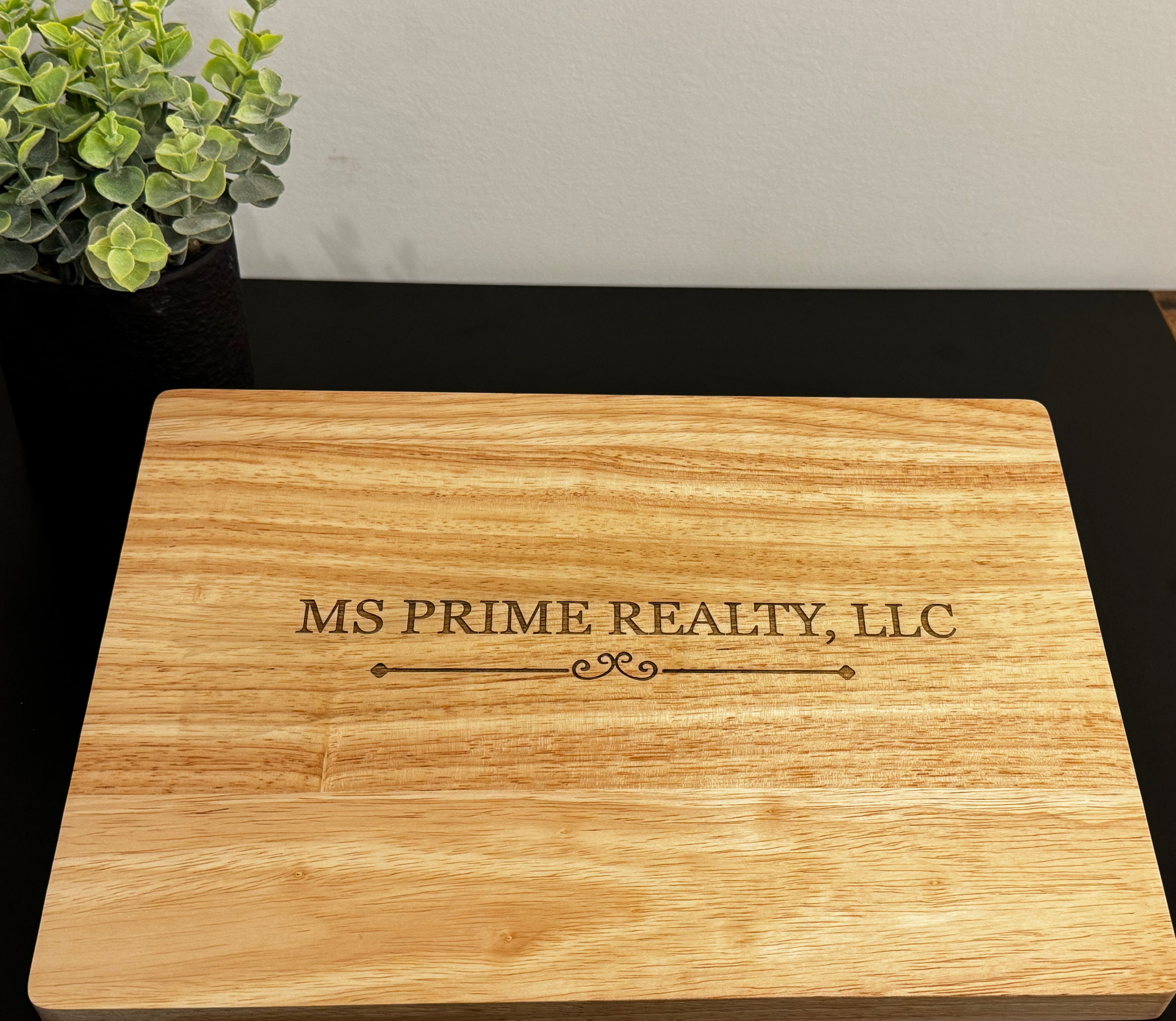 A rectangular wooden cutting board with natural grain, featuring the engraved text 'MS PRIME REALTY, LLC' in the center, resting on a black tabletop with a small potted plant in the background.