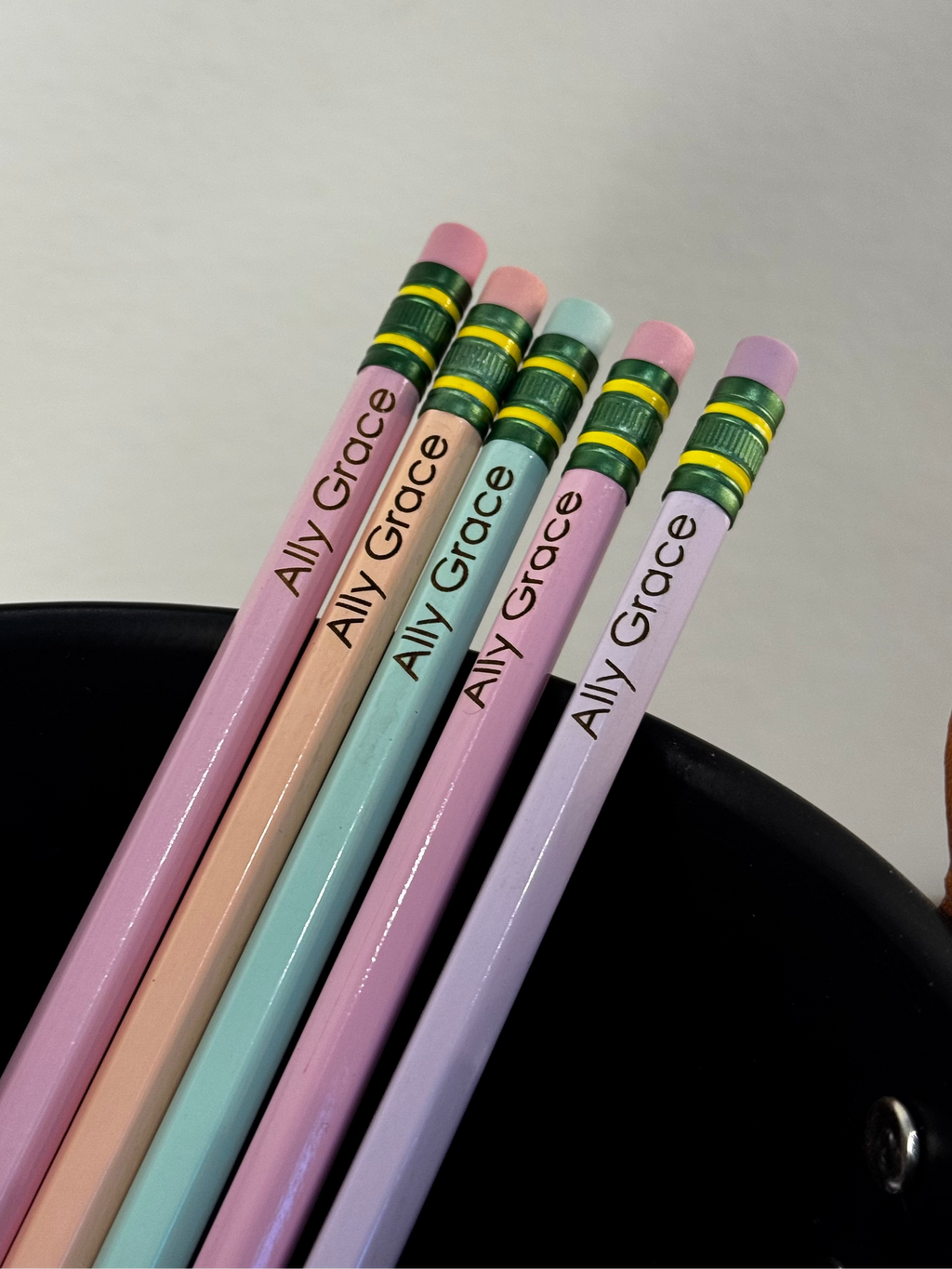 Five pastel-colored pencils with the name 'Alli Grace' printed on them, resting in a black cup against a light gray background.