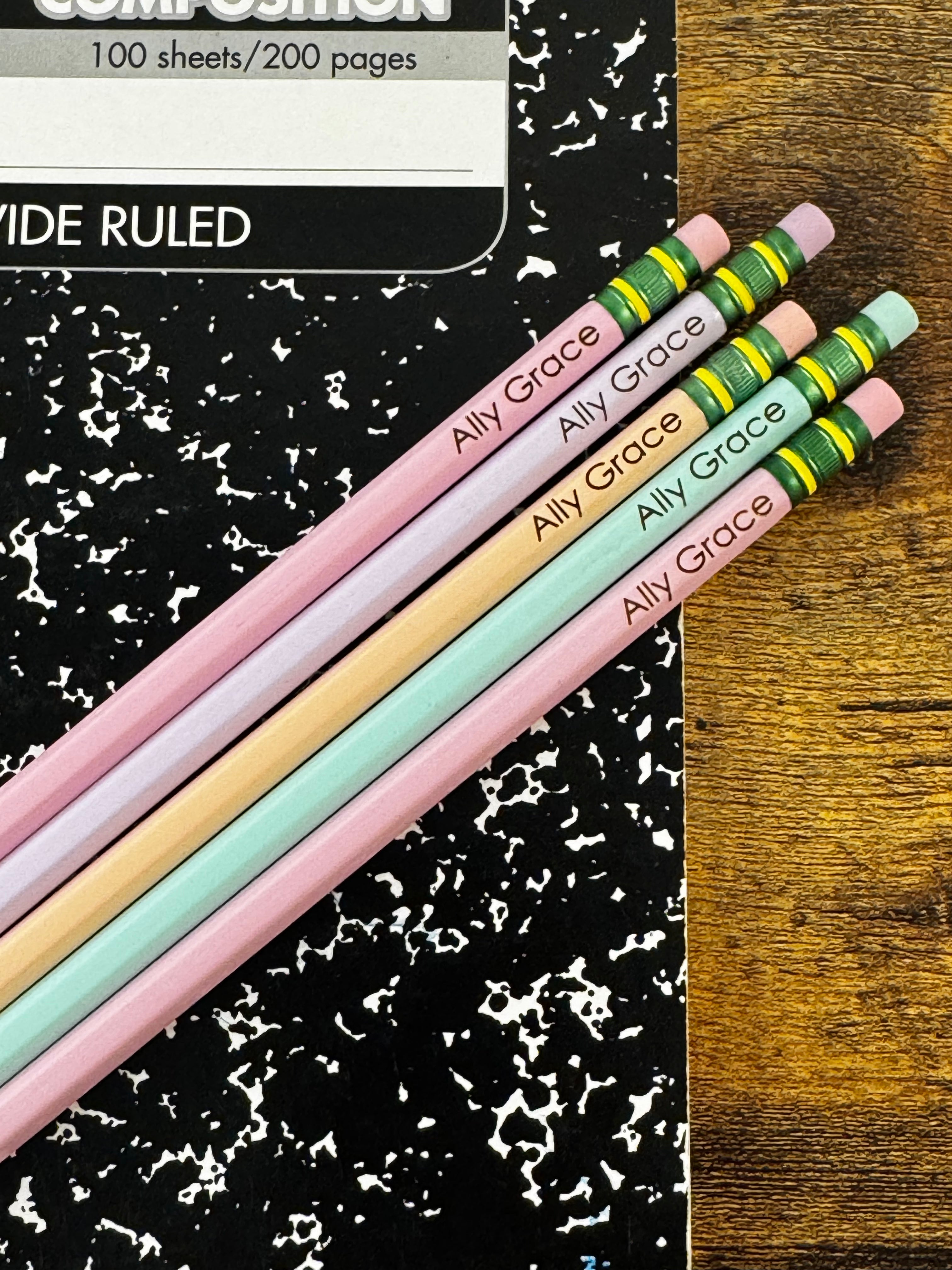 Four personalized wooden pencils in pastel pink, peach, mint green, and light blue, printed with the name 'Ally Grace' and featuring green and yellow ferrules, arranged diagonally on a black and white speckled composition notebook next to a wooden surface.