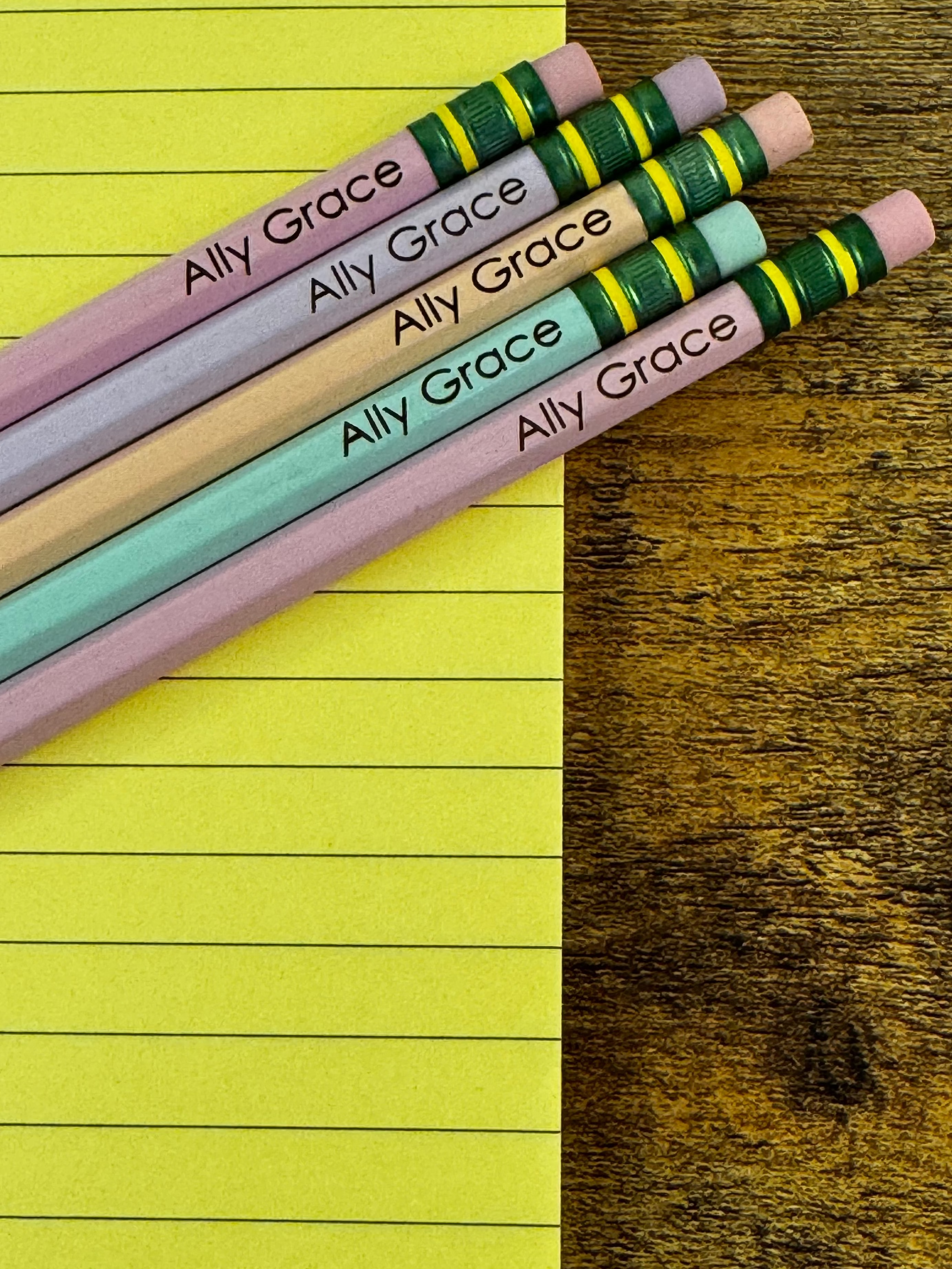 Four personalized wooden pencils with pink erasers and green ferrules, printed with the name 'Ally Grace', arranged diagonally on a split background of yellow lined paper and a dark wood surface.