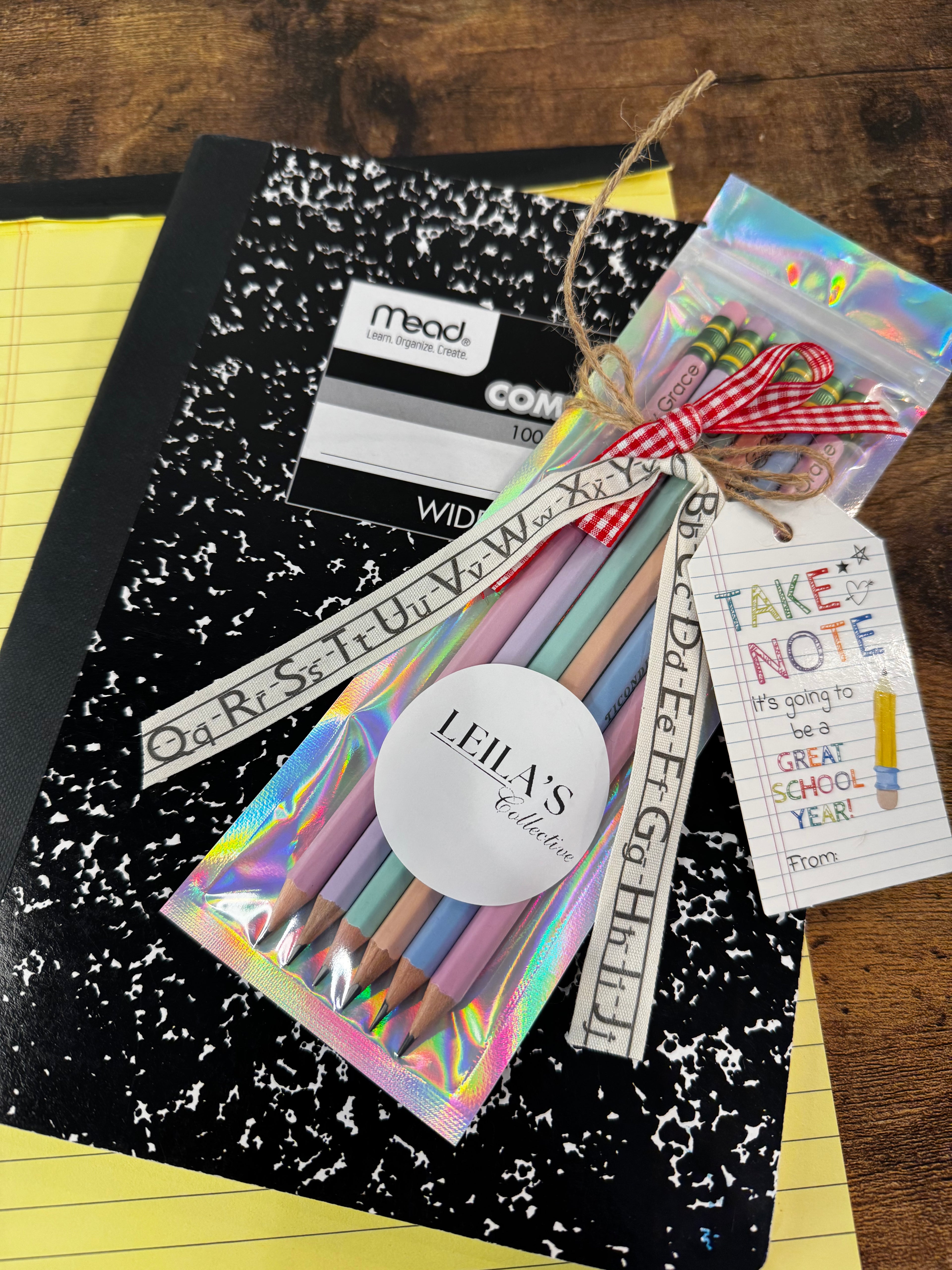 A school supply gift set featuring a black and white speckled notebook, a pack of pastel colored pencils in an iridescent sleeve, a red gingham ribbon, and a 'TAKE NOTE' tag, arranged on a wooden table.