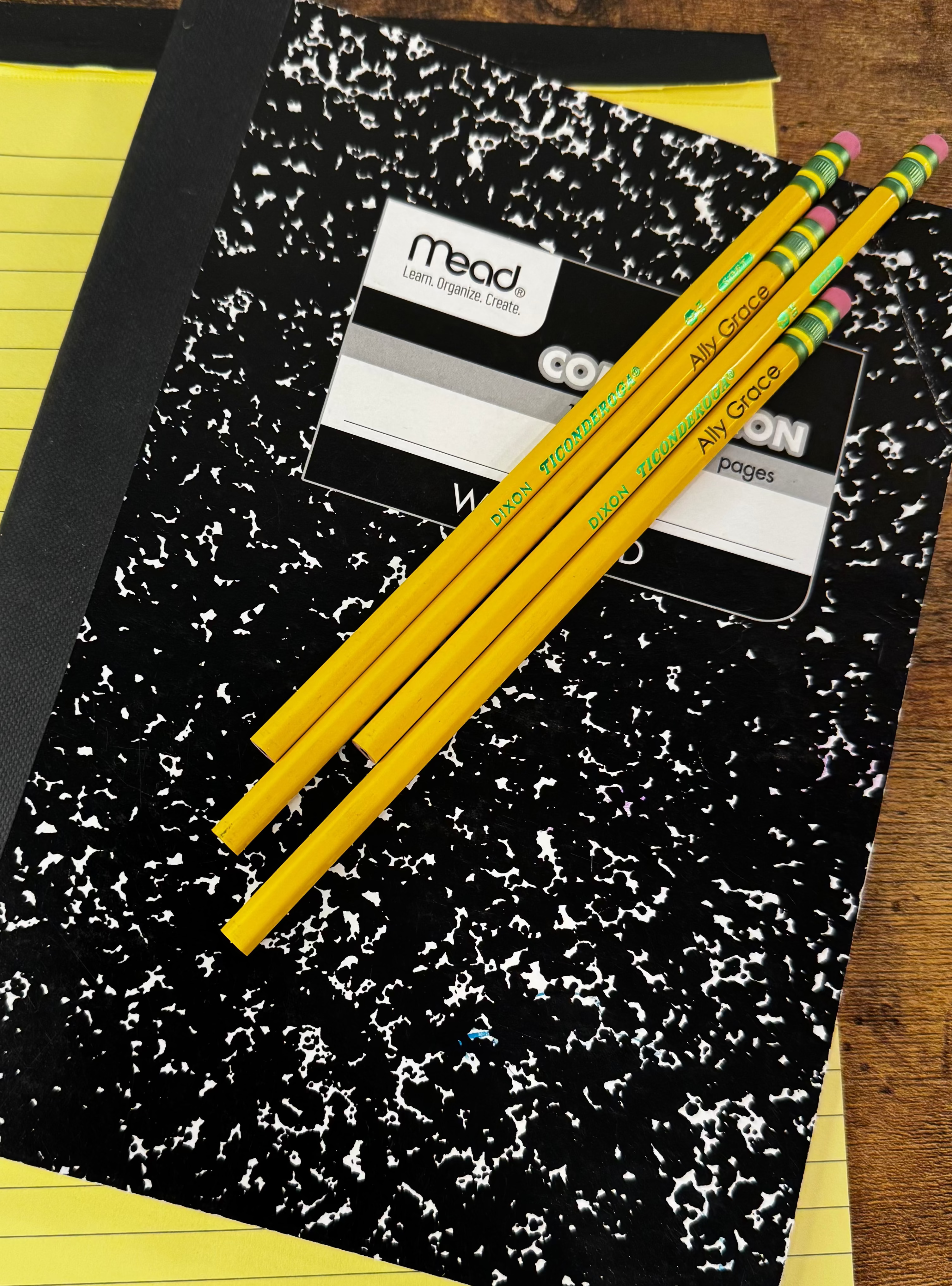 A black and white speckled composition notebook with a 'Mead' brand label, placed on top of a yellow ruled notepad on a wooden table, with three yellow pencils resting on the notebook.